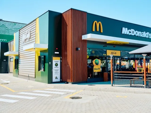 O clădire modernă cu fațadă verde, maro și galbenă prezintă un semn McDonald's deasupra unei intrări cu geamuri mari și o terasă metalică.