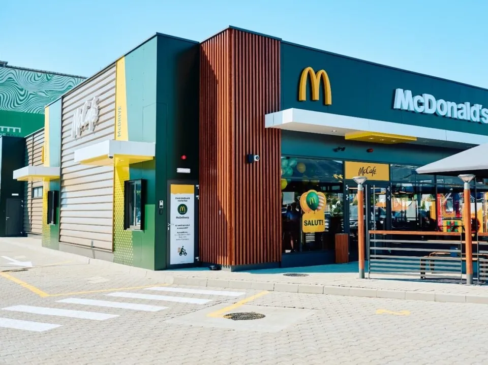O clădire modernă cu fațadă verde, maro și galbenă prezintă un semn McDonald's deasupra unei intrări cu geamuri mari și o terasă metalică.