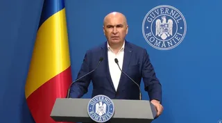 Un bărbat în costum bleumarin și camasă albă vorbește la un pupitru cu sigla Guvernului României, în fața unei fundal albastre cu steagul României.