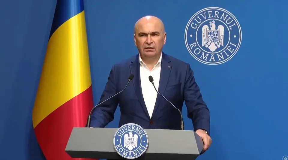 Un bărbat cu sacou albastru și cămașă albă stă în spatele unui podium gri cu emblema Guvernului României, în fața unui fundal albastru și a unui steag.