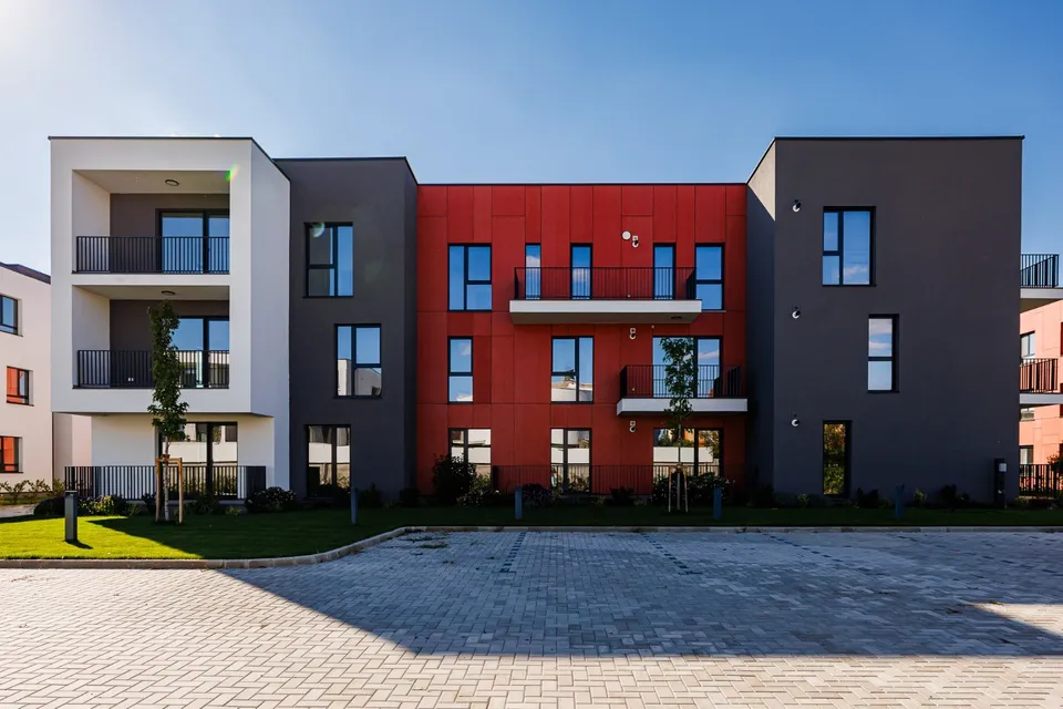 Un complex de clădiri moderne cu fațade gri, albe și roșii, cu ferestre și balcoane, situat în fața unui teren pavat.