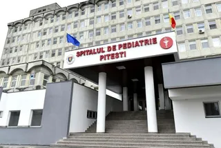 Intrarea unui spital este marcată de un panou alb cu textul roșu și un simbol medical, susținut de coloane albe lângă scări.