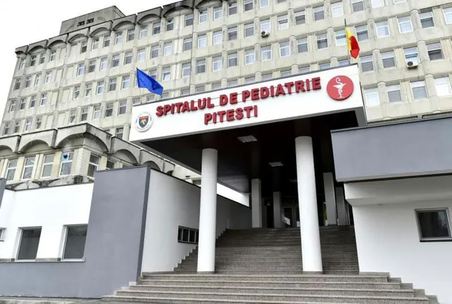 Intrarea unui spital este marcată de un panou alb cu textul roșu și un simbol medical, susținut de coloane albe lângă scări.