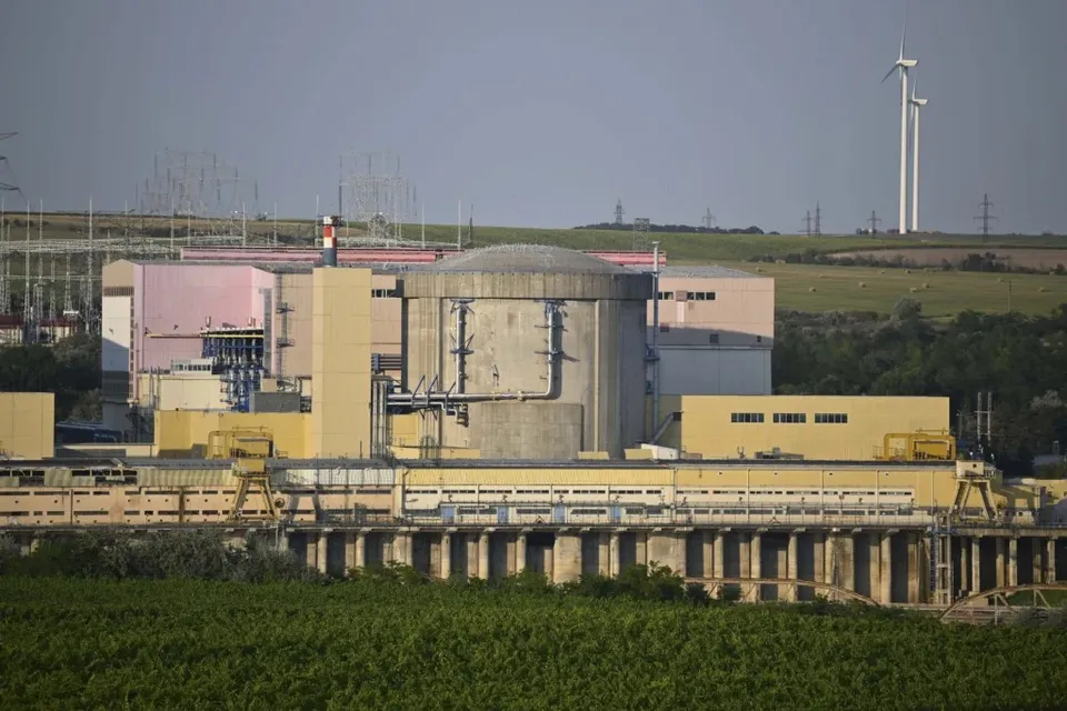 O clădire industrială mare cu secțiuni galbene și roz este situată lângă o structură cilindrică gri sub un cer senin.