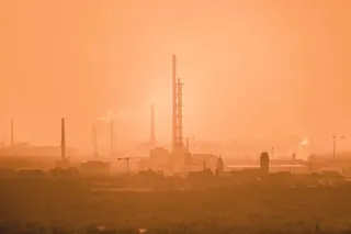 Siluete de coloane înalte și clădiri industriale sunt vizibile sub un cer de culoare portocalie, într-o atmosferă densă și încețoșată.