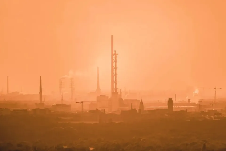 Siluete de coloane înalte și clădiri industriale sunt vizibile sub un cer de culoare portocalie, într-o atmosferă densă și încețoșată.