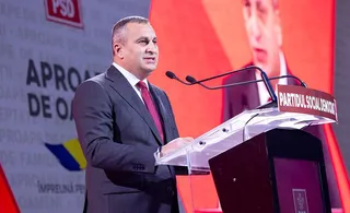 Un bărbat într-un costum gri și cravată roșie vorbește la un podium transparent în fața unui panou roșu și alb.