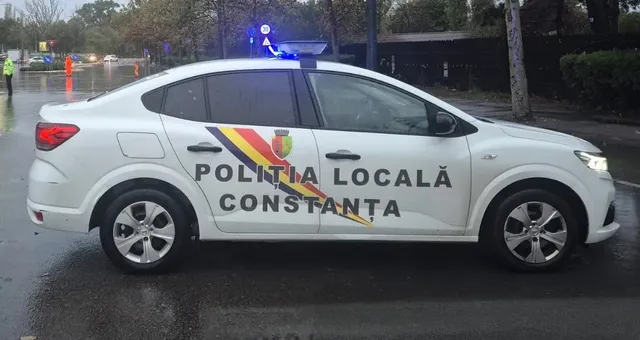 Un autovehicul alb cu inscripția Poliția Locală Constanța și un tricolor pe lateral este parcat pe un drum umed sub lumină albastră.