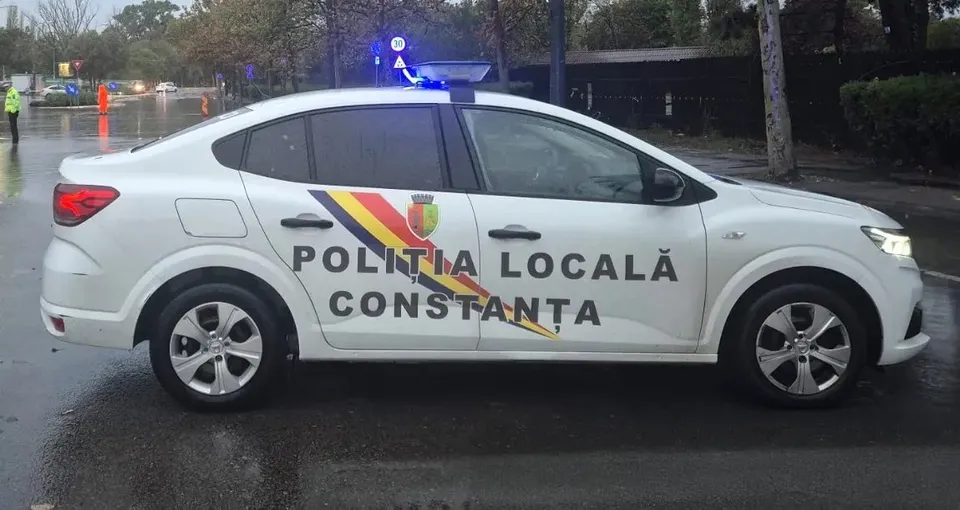 Un autovehicul alb cu inscripția Poliția Locală Constanța și un tricolor pe lateral este parcat pe un drum umed sub lumină albastră.