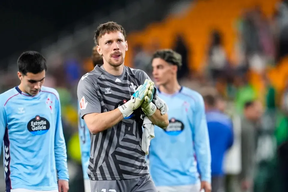 Un fotbalist cu mâniile în mănuși de portar, purtând un echipament gri cu dungi negre, stă pe un teren de fotbal lângă alți doi jucători.