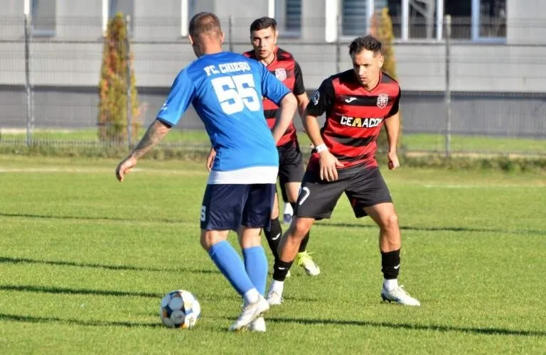 Trei jucători de fotbal sunt pe un teren de gazon verde, unul purtând un tricou albastru cu numărul cincizeci și cinci și celălalt un tricou roșu.