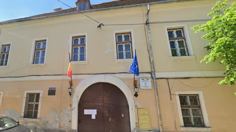 O clădire cu două etaje prezintă ferestre dreptunghiulare, un arc de intrare maro și două steaguri suspendate pe fațadă.