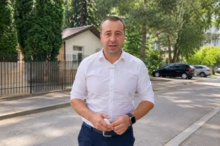 Un bărbat cu cămașă albă și pantaloni închiși stă în picioare pe un trotuar lângă un gard metalic și mașini parcate.