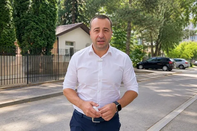 Un bărbat cu cămașă albă și pantaloni închiși stă în picioare pe un trotuar lângă un gard metalic și mașini parcate.