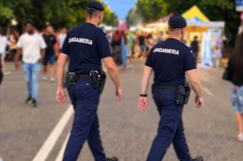 Doi jandarmi în uniforme albastre cu inscripția JANDARMERIA merg pe un drum asfaltat printre mulțime.