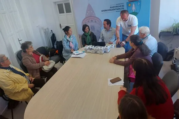 Oamenii stau la o masă lungă de lemn în jurul căreia se află cutii mici albe și un bărbat în tricou alb aplecat.