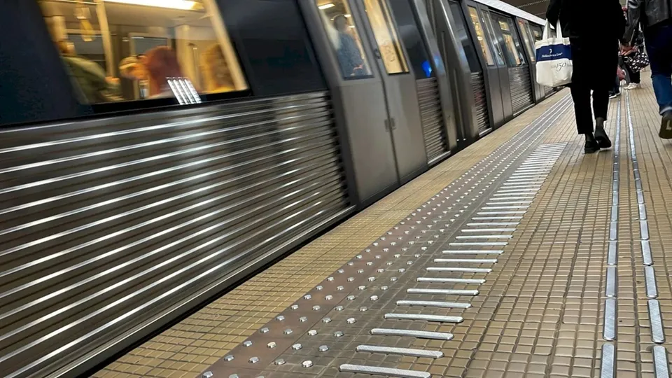 Un tren de metrou gri stă lângă un peron de stație, în timp ce o persoană în haine negre merge pe marginea liniei de siguranță.
