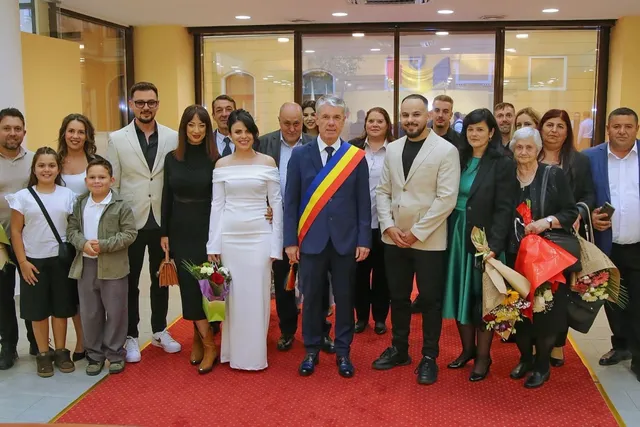 Un grup de persoane stă în rând pe un covor roșu în fața unei ferestre mari, purtând haine de sărbătoare și ținând buchete de flori.