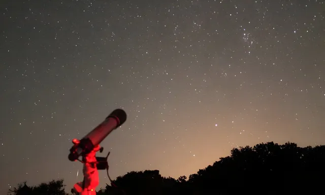 Un telescop negru este poziționat pe un trepied sub un cer nocturn plin de stele, deasupra unei siluete de copaci întunecați.