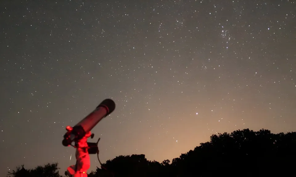 Un telescop negru este poziționat pe un trepied sub un cer nocturn plin de stele, deasupra unei siluete de copaci întunecați.