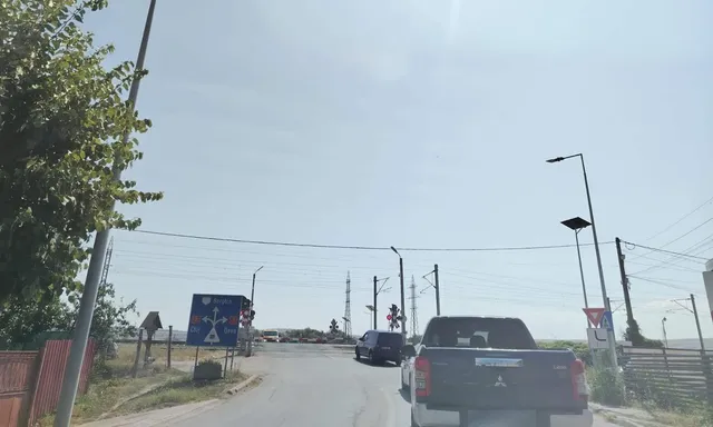 Un pick-up gri și un autoturism negru circulă pe un drum asfaltat sub un cer senin, lângă un semn de trecere la nivel și stâlpi de iluminat.