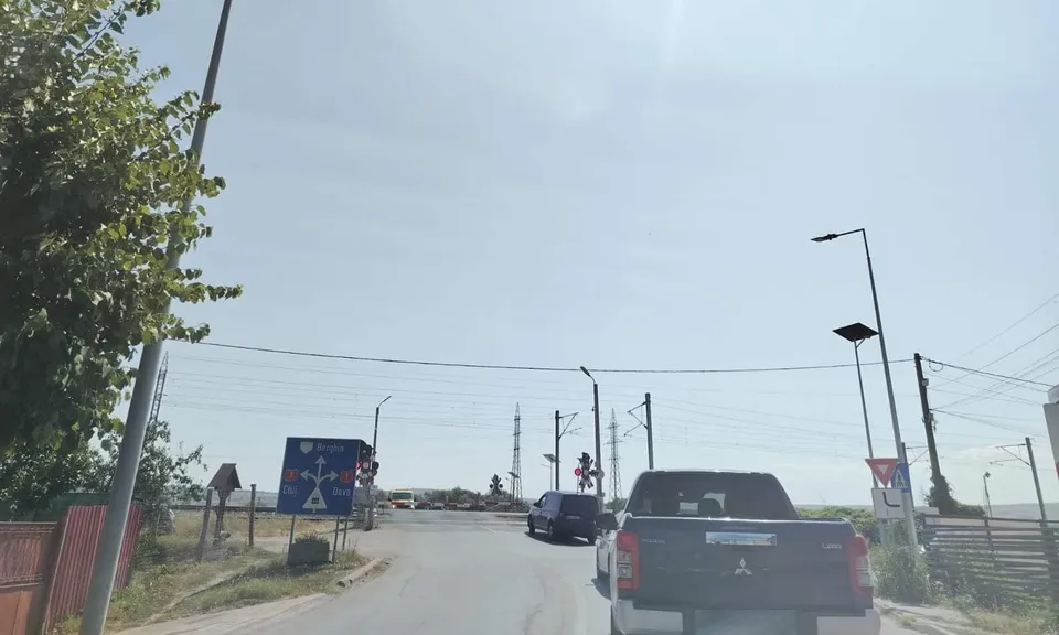 Un pick-up gri și un autoturism negru circulă pe un drum asfaltat sub un cer senin, lângă un semn de trecere la nivel și stâlpi de iluminat.