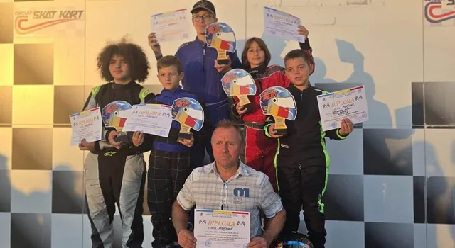 Șase copii și un adult stau în față, ținând diplome și trofee pe un fundal cu modele de pătrate alb-negre.