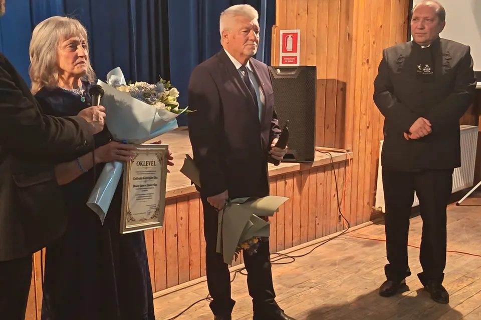 Un bărbat într-un costum închis și o femeie cu o rochie închisă țin buchete de flori și un diplo pe un decor din lemn.