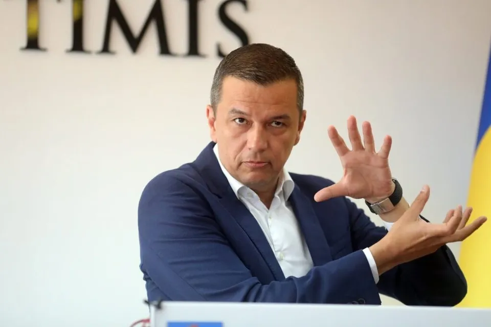 Un bărbat cu sacou albastru și cămașă albă stă la o masă cu mâinile ridicate în poziție de gesticulație în fața unui fundal alb cu litere negre.