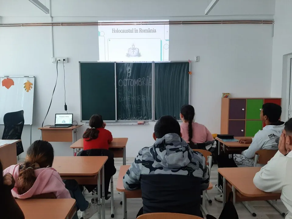 Elevi stau la bănci într-o sală de clasă privind un ecran de proiecție care afișează un text pe un fundal alb.