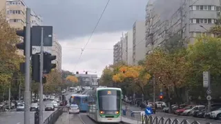Un tramvai verde și alb circulă pe șine într-o zonă urbană cu clădiri înalte, mașini parcate și copaci cu frunziș galben sub un cer înnorat.