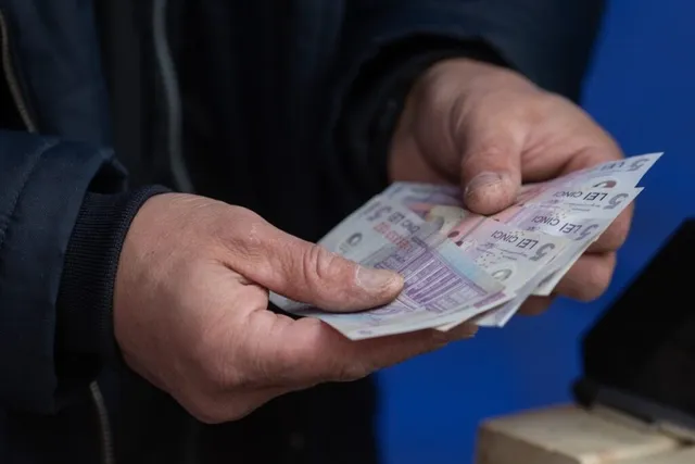 Două mâini țin mai multe bancnote de cinci lei deasupra unei suprafețe de culoare albă.