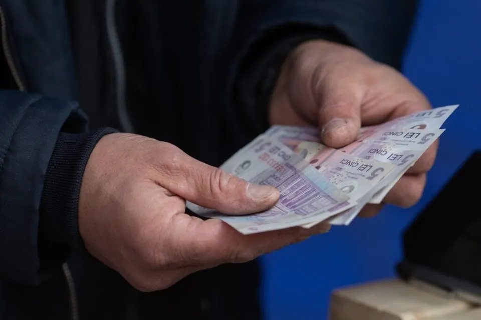 Două mâini țin mai multe bancnote de cinci lei deasupra unei suprafețe de culoare albă.
