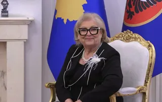 O femeie cu ochelari și colier de perle stă pe un scaun decorat cu auriu în fața unor steaguri albastre.