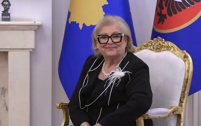 O femeie cu ochelari și colier de perle stă pe un scaun decorat cu auriu în fața unor steaguri albastre.