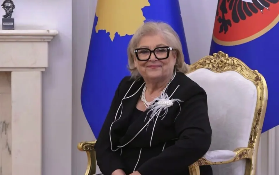 O femeie cu ochelari și colier de perle stă pe un scaun decorat cu auriu în fața unor steaguri albastre.