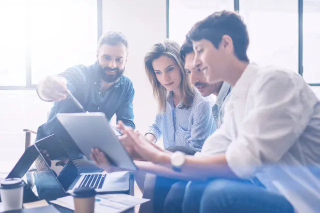 Un grup de patru persoane stă la o masă și privește un dispozitiv digital plat în timp ce unul dintre ei ține un stilou.