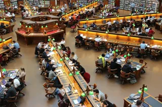 Oamenii stau la mese lungi de lucru în interiorul unei biblioteci mari, folosind laptopuri și lămpi de birou sub o lumină caldă.