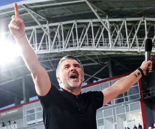 Un bărbat cu tricou negru stă cu brațele ridicate, ținând un microfon în mâna stângă, sub o structură metalică de stadion.