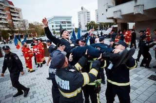 Un grup de bărți în uniforme de serviciu negre cu benzi galbene îi ridică pe doi bărți în aer pe o piață pavată.
