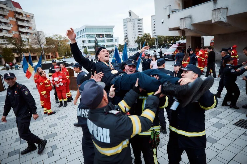 Un grup de bărți în uniforme de serviciu negre cu benzi galbene îi ridică pe doi bărți în aer pe o piață pavată.
