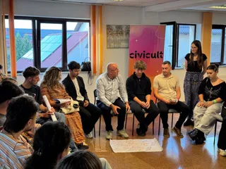 Un grup de persoane stă așezat pe scaune într-o încăpere cu ferestre mari, privind spre un bărbat în cămașă albă și pantaloni închiși.