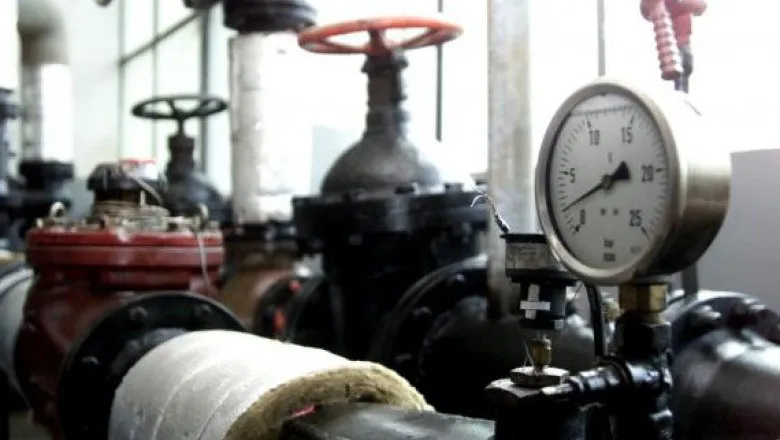 Un manometru cu cadran alb afișează valori numerice, fiind montat pe o conductă metalică lângă o supapă cu roată roșie într-o instalație industrială.