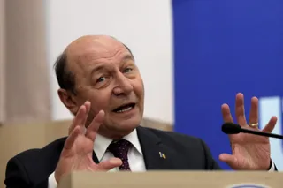Un bărbat cu părul rare și costum închis, cămașă albă și cravată mov, stă la un microfon cu mâinile ridicate.