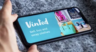 O mână ține un smartphone afișând logo-ul Vinted și produse de îmbrăcăminte pe un fundal texturat de stofă.