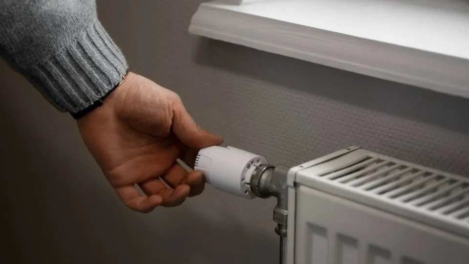 O mână întinde degetul spre un termostat alb montat pe un radiator de culoare albă sub un pervaz.