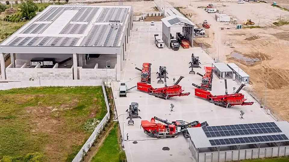 O vedere aeriană prezintă o clădire cu panouri solare pe acoperiș și diverse utilaje industriale de culoare roșie parcate pe o suprafață betonată.