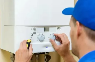 Un tehnician cu o șapcă albastră reglează butoanele unei centrale termice albe folosind o șurubelniță.