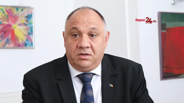 Un bărbat cu părul scurt și costum negru, cămașă albă și cravată albastră, poartă un pin pe reverul sacoului în fața unui fundal alb.