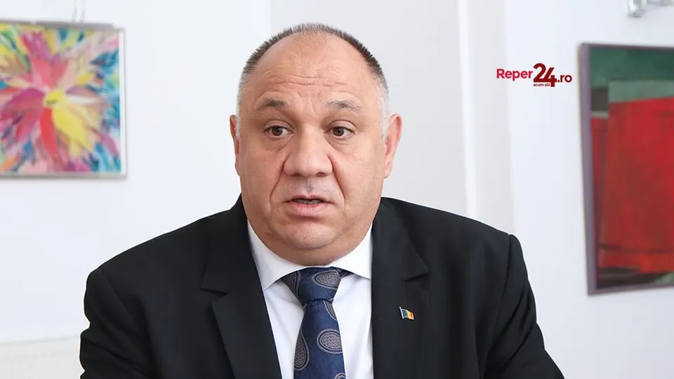 Un bărbat cu părul scurt și costum negru, cămașă albă și cravată albastră, poartă un pin pe reverul sacoului în fața unui fundal alb.